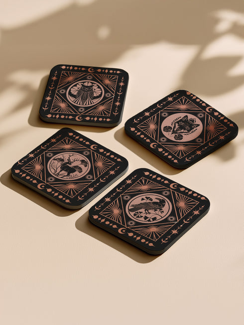 A set of four black, high-gloss square coasters featuring intricate rose-gold metallic illustrations of an owl, raven, stag, and wolf framed by celestial and botanical motifs. Made from durable masonite hardboard with cork backing for slip-resistant protection.